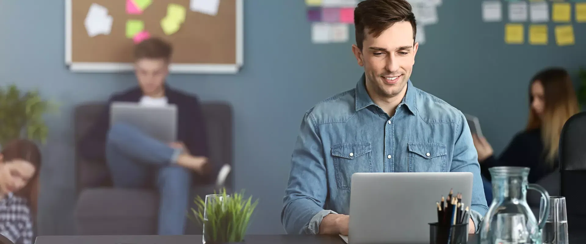 Ein Mann im Jeanshemd verwendet in einer geschäftigen Büroumgebung einen Laptop zur Marketingunterstützung, während im Hintergrund Kollegen arbeiten.
