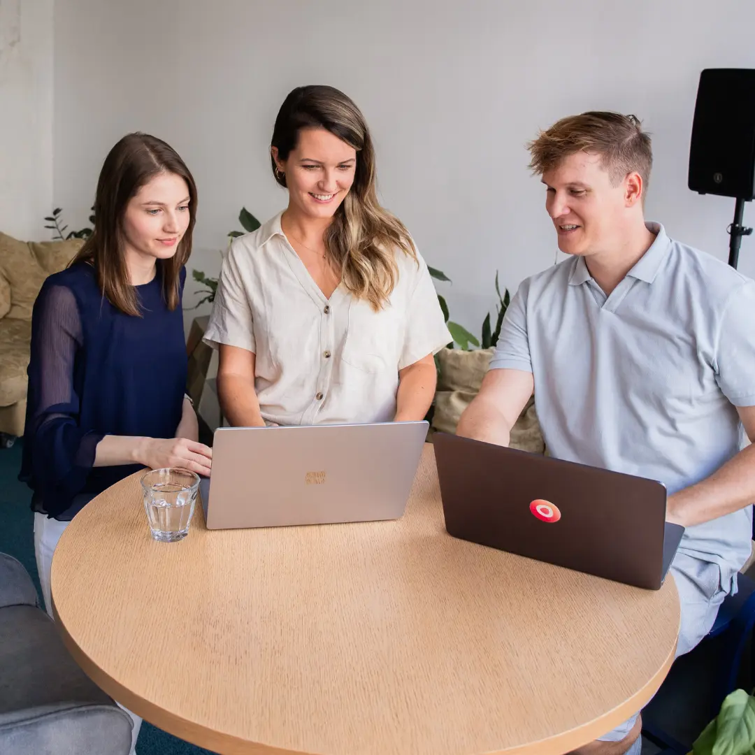 Drei Personen aus dem Einfachmarketing-Team arbeiten an Laptops an einem runden Tisch in einem modernen Büroraum.
