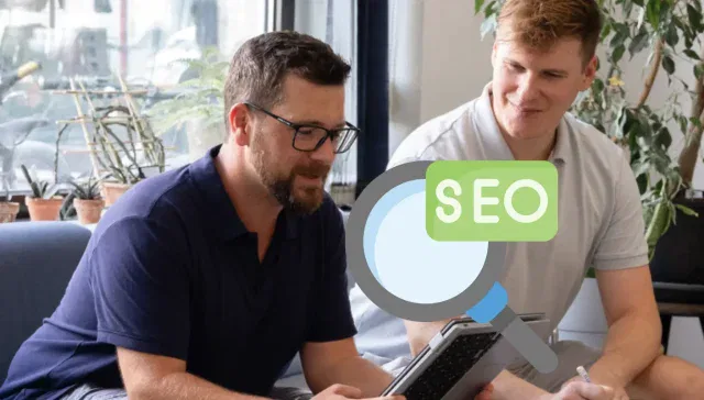 Zwei Personen sitzen auf einem Sofa und diskutieren über SEO. Eine Person hält ein Tablet in der Hand. Über das Bild ist eine Grafik einer Lupe mit der Aufschrift „SEO“ gelegt. Dies unterstreicht, wie wichtig es ist, die Suchabsicht zu verstehen, um Suchabsichtsstrategien effektiv zu verbessern.