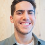 Ein Mann mit dunklem Haar und Bart, der den Charme des Einfachmarketing-Teams versprüht, lächelt breit. Er trägt ein grünes Hemd über einem weißen T-Shirt und steht selbstbewusst vor einem hellen Holzhintergrund.