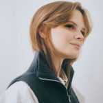 Ein Mitglied des Einfachmarketing-Teams mit langen Haaren und einem leichten Lächeln, das eine dunkle Weste mit Reißverschluss über einem weißen Hemd trägt, blickt vor einem schlichten Hintergrund nachdenklich zur Seite.