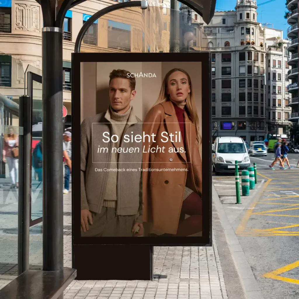 Eine Straßenszene in einer Stadt zeigt eine Bushaltestellenwerbung, die einen Mann und eine Frau in stilvoller Kleidung mit deutschem Text zeigt, die für die Modemarke Schanda als automatisch gespeicherter Entwurf werben.