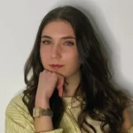 Eine Frau mit langen braunen Haaren, gelb gestreiftem Hemd und goldener Uhr posiert mit auf die Hand gestütztem Kinn und verkörpert so den sympathischen Stil des Einfachmarketing-Teams vor einem schlichten weißen Hintergrund.