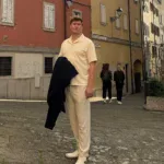 Leonhard Pleschberger in leichter Kleidung steht auf einem kopfsteingepflasterten Platz, umgeben von bunten Gebäuden unter einem bewölkten Himmel. Im Hintergrund sind ein paar Menschen zu sehen.