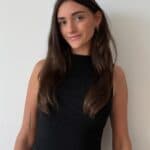 Eine Frau mit langen braunen Haaren, die zum Einfachmarketing-Team gehört, steht in einem ärmellosen schwarzen Oberteil vor einer schlichten weißen Wand und lächelt leicht. Sie hat eine Tätowierung auf ihrem linken Unterarm und trägt goldene Ohrringe.