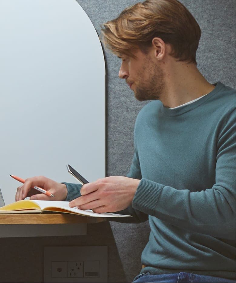 Ein Mann in einem blaugrünen Pullover sitzt an einem Schreibtisch und schreibt mit einem Bleistift in ein Notizbuch, in der anderen Hand hält er ein Telefon.