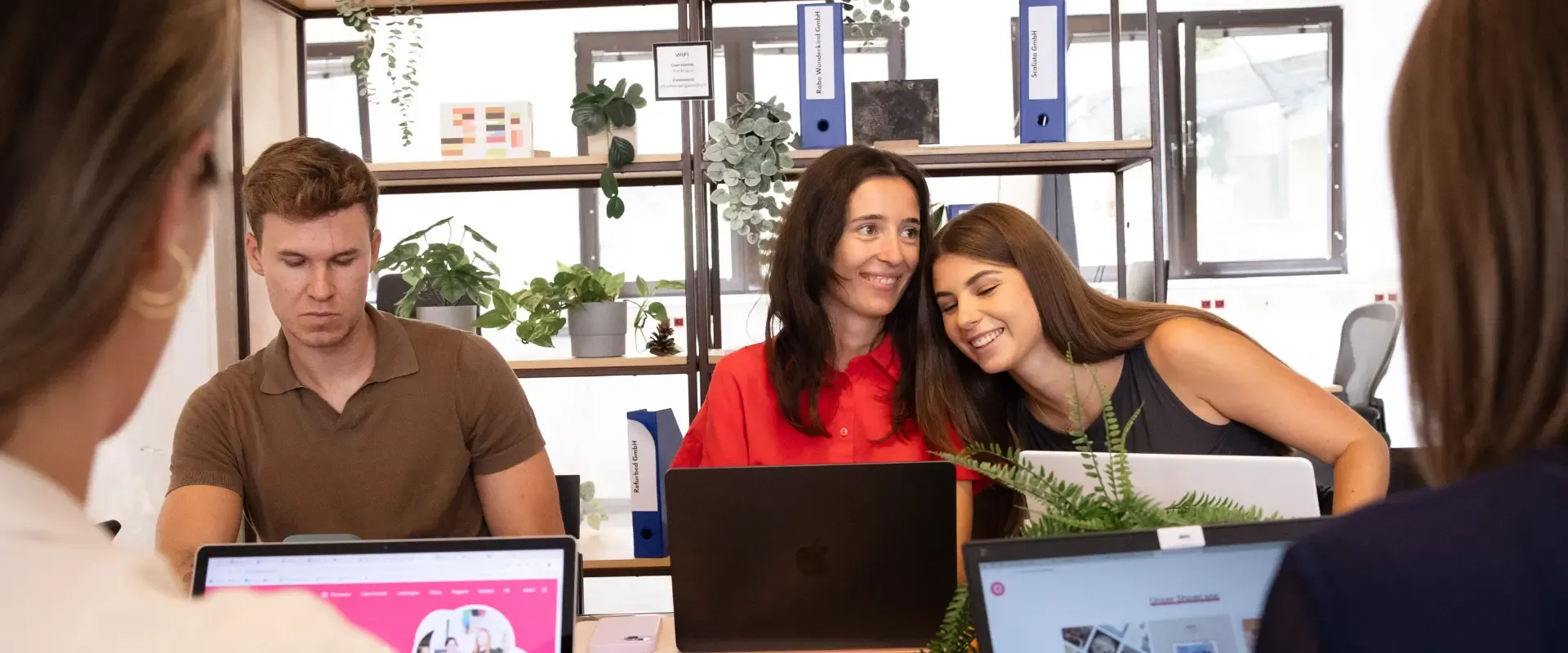 Zwei Frauen sitzen zusammen an einem Schreibtisch, eine in einem roten Hemd und die andere in einem grauen, und teilen einen Moment über Social-Media-Tools. Ein Mann in einem braunen Hemd arbeitet an einem separaten Schreibtisch. Im Hintergrund sind verschiedene Laptops und Pflanzen zu sehen.