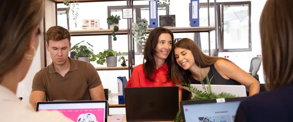 Zwei Frauen sitzen an einem Tisch und arbeiten an Laptops. Eine lehnt ihren Kopf an die Schulter der anderen und erkundet wahrscheinlich Social-Media-Tools. Ein Mann arbeitet ebenfalls in der Nähe an einem Laptop. Im Hintergrund sind Büropflanzen und Bücherregale zu sehen.