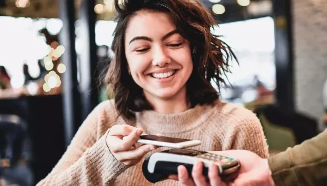 Eine lächelnde Frau benutzt ihr Smartphone, um in einem belebten Café an einem Kartenleser kontaktlos zu bezahlen.