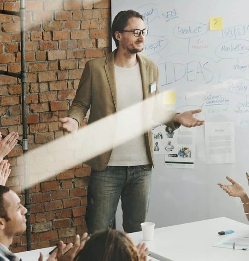 Ein Mann mit Brille und beigem Blazer steht vor einem Whiteboard mit Brainstorming-Notizen und hält eine Präsentation vor einer Gruppe klatschender Menschen in einem Raum mit einer Ziegelwand.