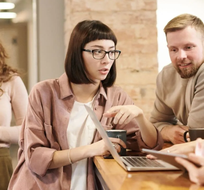 Eine Frau mit Brille und ein Mann schauen auf einen Laptop auf einem Holztisch. Die Frau zeigt auf den Bildschirm und hält dabei eine Kaffeetasse in der Hand. Im Hintergrund ist eine weitere Person teilweise zu sehen.