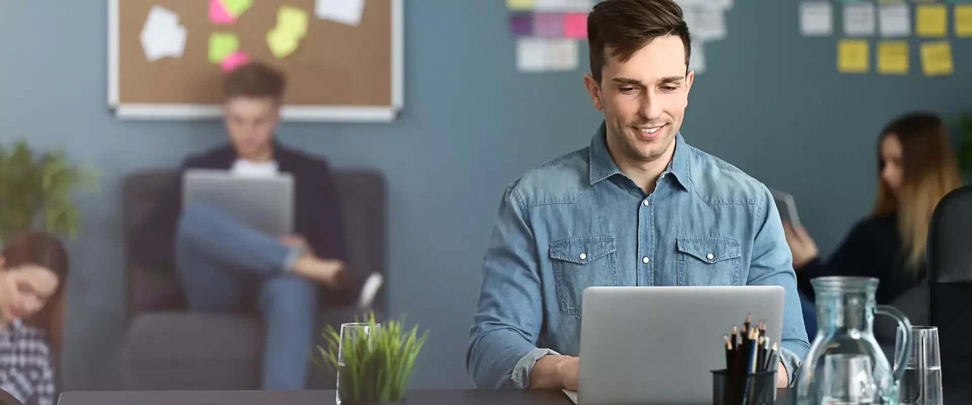 Ein Mann im Jeanshemd verwendet in einer geschäftigen Büroumgebung einen Laptop zur Marketingunterstützung, während im Hintergrund Kollegen arbeiten.