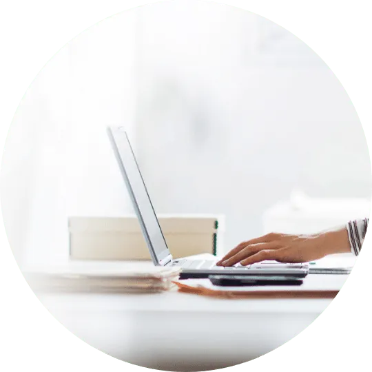 Eine Person tippt auf einem Laptop an einem Schreibtisch, neben ihr ein Smartphone und Papierkram.