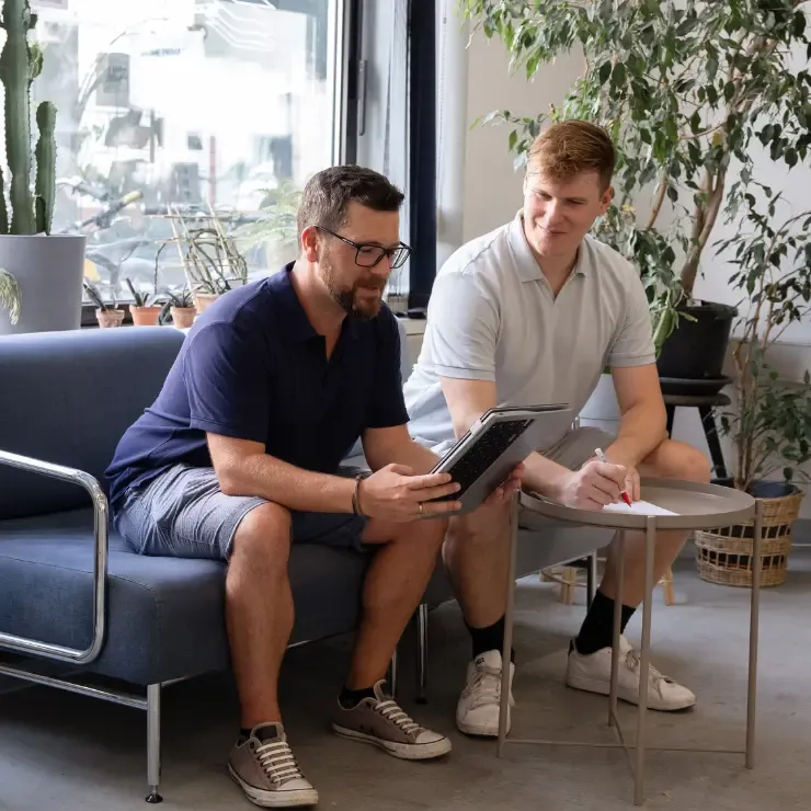 Zwei Männer sitzen auf einem blauen Sofa. Einer hält ein Tablet und zeigt es dem anderen, der auf einem Notizblock schreibt. Sie befinden sich in einem gut beleuchteten Raum mit Pflanzen am Fenster und arbeiten gemeinsam an ihren Marketing-Abo-Strategien.