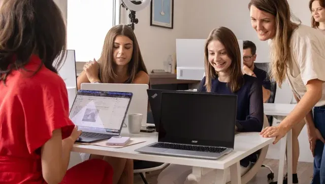 Eine Gruppe von Marketingberatern arbeitet in einer Büroumgebung zusammen und sitzt an einem Tisch an Laptops. Im Hintergrund sind ein Whiteboard und andere Bürogeräte zu sehen.