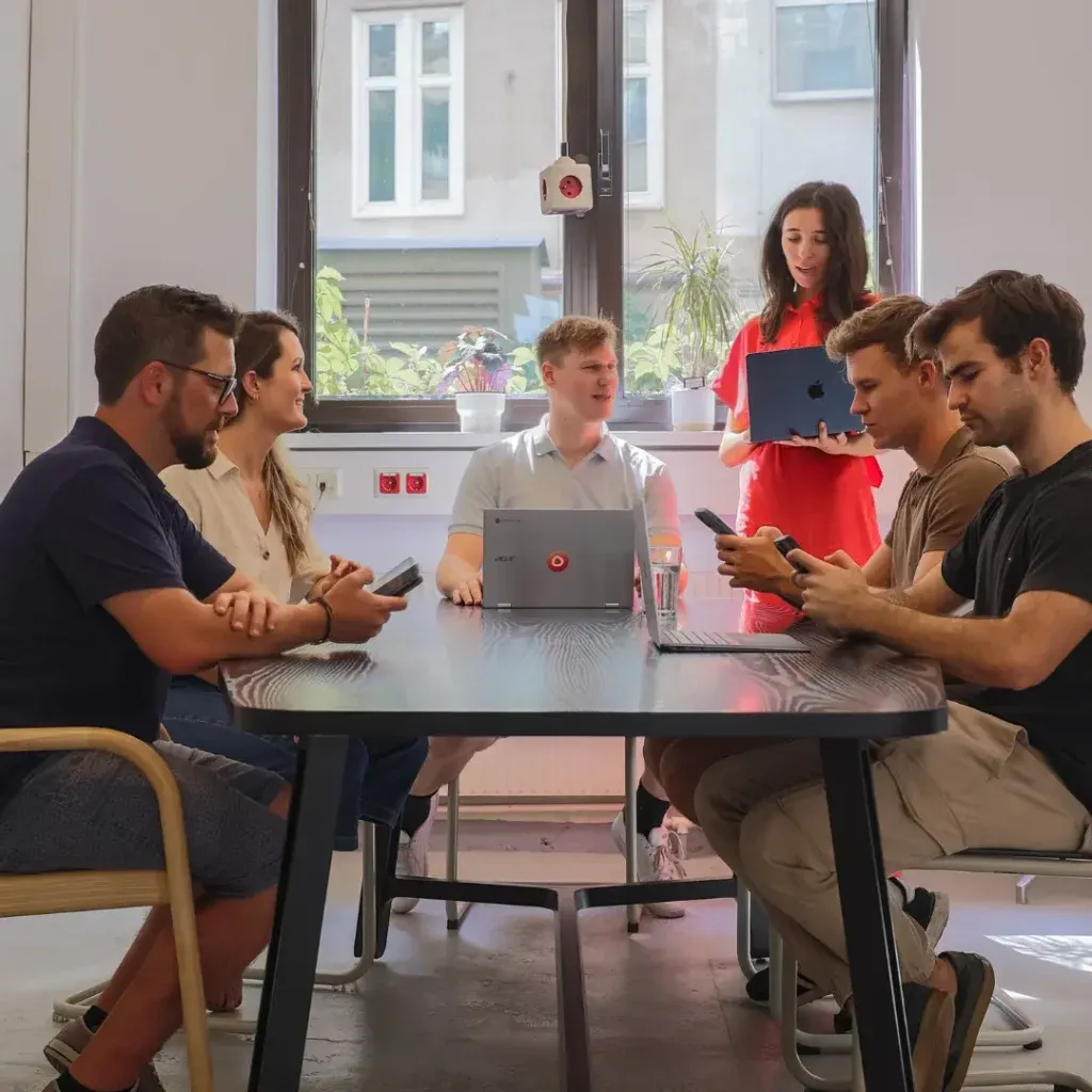 Sechs Mitglieder des Einfachmarketing-Teams sind in einem Büro um einen Tisch versammelt; einige sitzen mit ihren Telefonen, während einer mit einem Laptop in der Hand dasteht und interagiert.