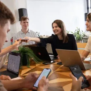 Menschen in einem Büro sitzen mit Laptops und Telefonen um einen Holztisch. Eine Frau in der Mitte reicht einem Mann einen Gegenstand. Der Teamgeist des Einfachmarketing-Teams wird deutlich, da eine Topfpflanze und andere Geräte den Tisch schmücken.
