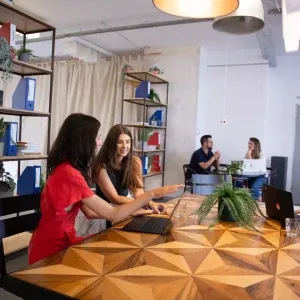 Zwei Frauen aus dem Einfachmarketing-Team arbeiten an Laptops an einem Holztisch in einem modernen Büro. Im Hintergrund unterhalten sich zwei weitere Personen. Geometrische Regale, Pflanzen und ein Vorhang schmücken den Raum.