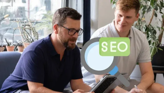 Zwei Personen sitzen auf einem Sofa und diskutieren über SEO. Eine Person hält ein Tablet in der Hand. Über das Bild ist eine Grafik einer Lupe mit der Aufschrift „SEO“ gelegt. Dies unterstreicht, wie wichtig es ist, die Suchabsicht zu verstehen, um Suchabsichtsstrategien effektiv zu verbessern.