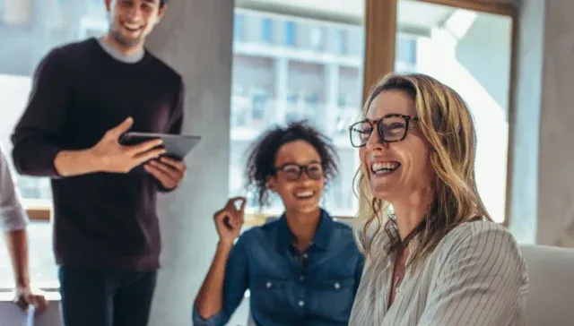 Vier Personen in legerer Businesskleidung unterhalten sich lächelnd in einer hellen Büroumgebung mit großen Fenstern über die Vorteile einer Marketing-Flatrate für Unternehmen im DACH-Raum.