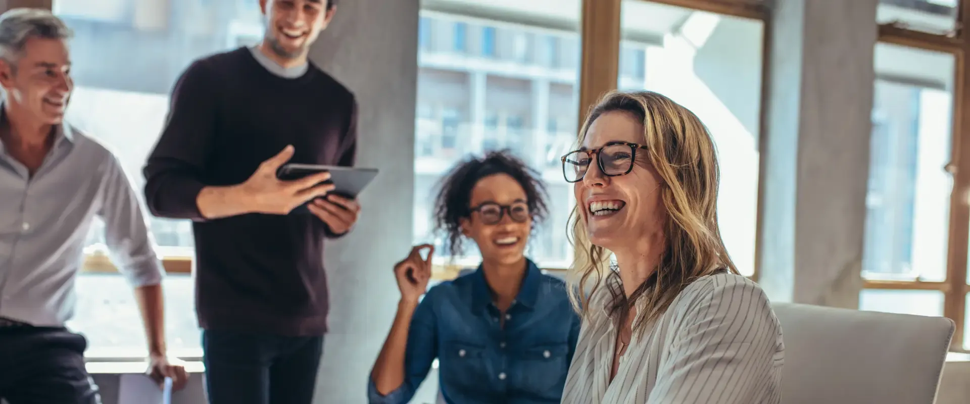 Vier Personen in legerer Businesskleidung unterhalten sich lächelnd in einer hellen Büroumgebung mit großen Fenstern über die Vorteile einer Marketing-Flatrate für Unternehmen im DACH-Raum.