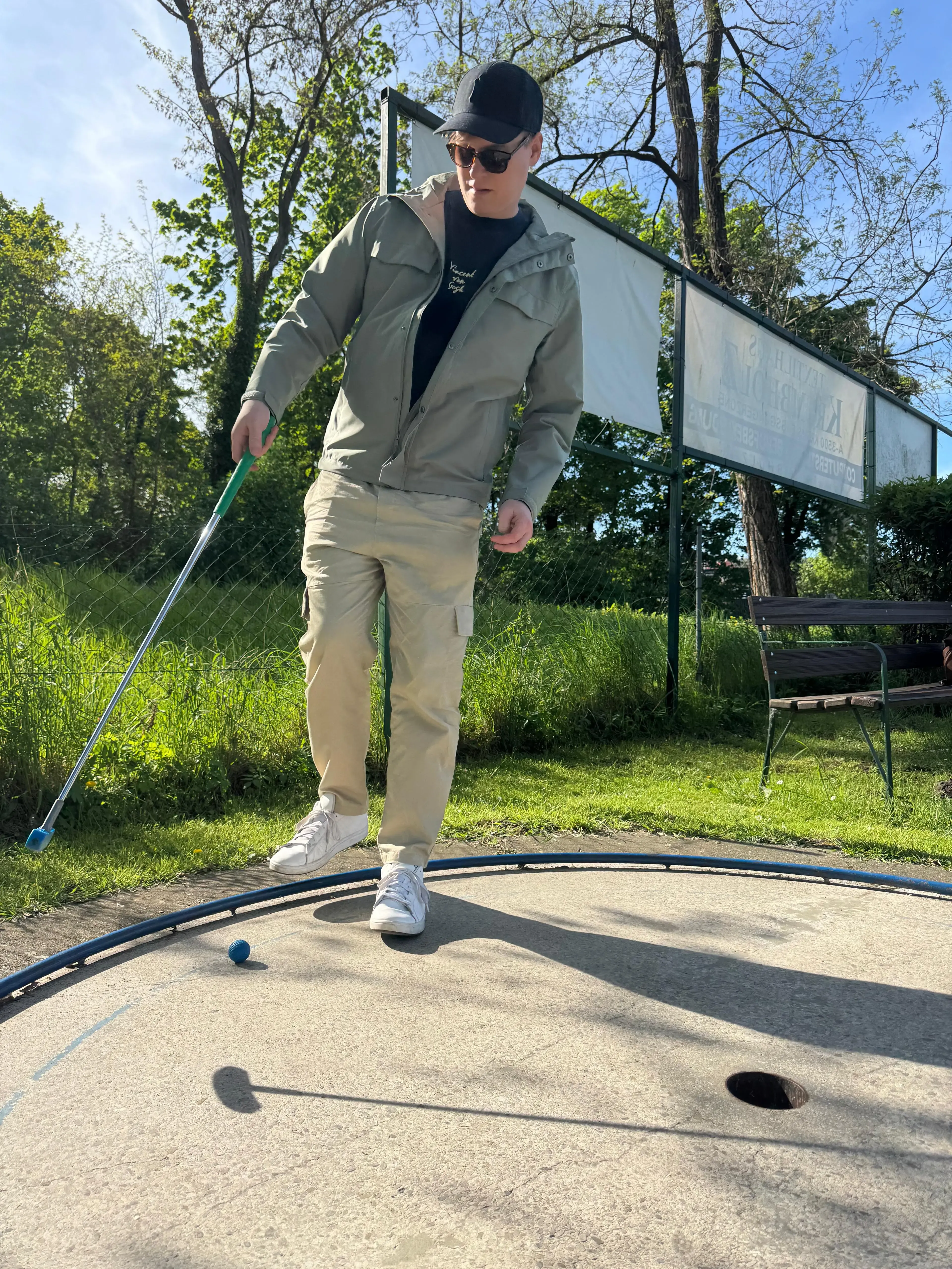 Leonhard Pleschberger, gekleidet in Freizeitkleidung, bereitet sich darauf vor, einen blauen Golfball in ein Loch auf einem Minigolfplatz im Freien zu schlagen.