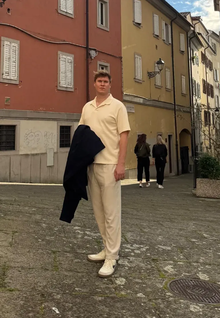 Leonhard Pleschberger in leichter Kleidung steht auf einem kopfsteingepflasterten Platz, umgeben von bunten Gebäuden unter einem bewölkten Himmel. Im Hintergrund sind ein paar Menschen zu sehen.
