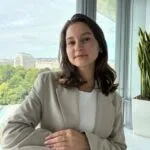 Eine Frau mit schulterlangen braunen Haaren in einem beigen Blazer und einem weißen Oberteil sitzt an einem weißen Tisch in der Nähe eines Fensters, mit Blick auf die Stadt und das Grün im Hintergrund - das fängt den professionellen Geist des Einfachmarketing-Teams ein.